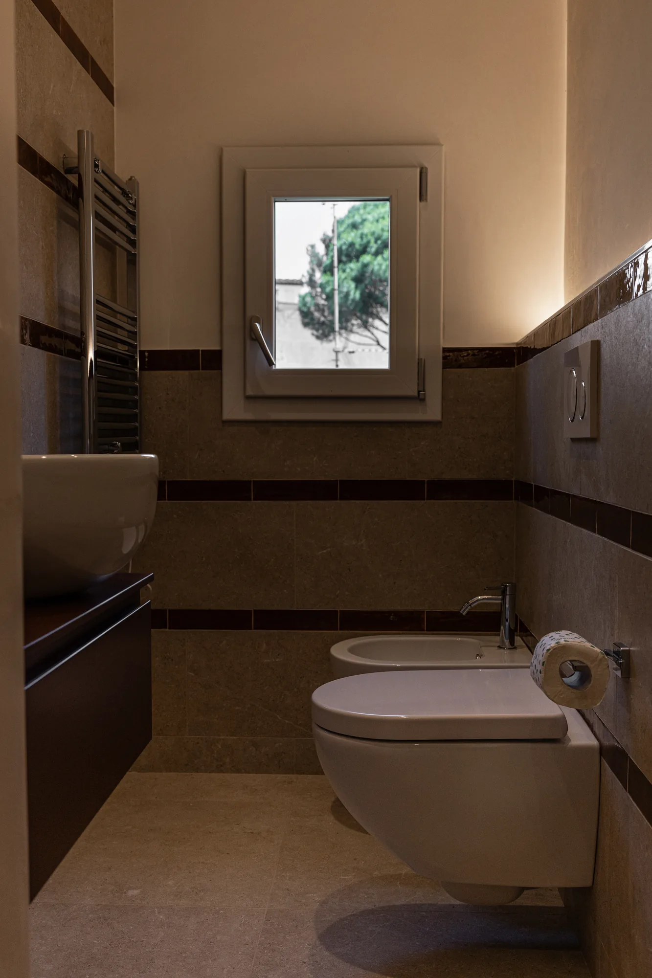 Narrow bathroom with wall-mounted toilet, bidet, window, and towel warmer