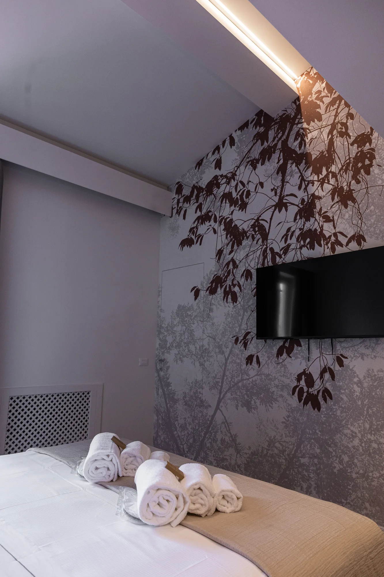 Bedroom with made bed, rolled towels, and wall decorated with leaf pattern and flat-screen TV