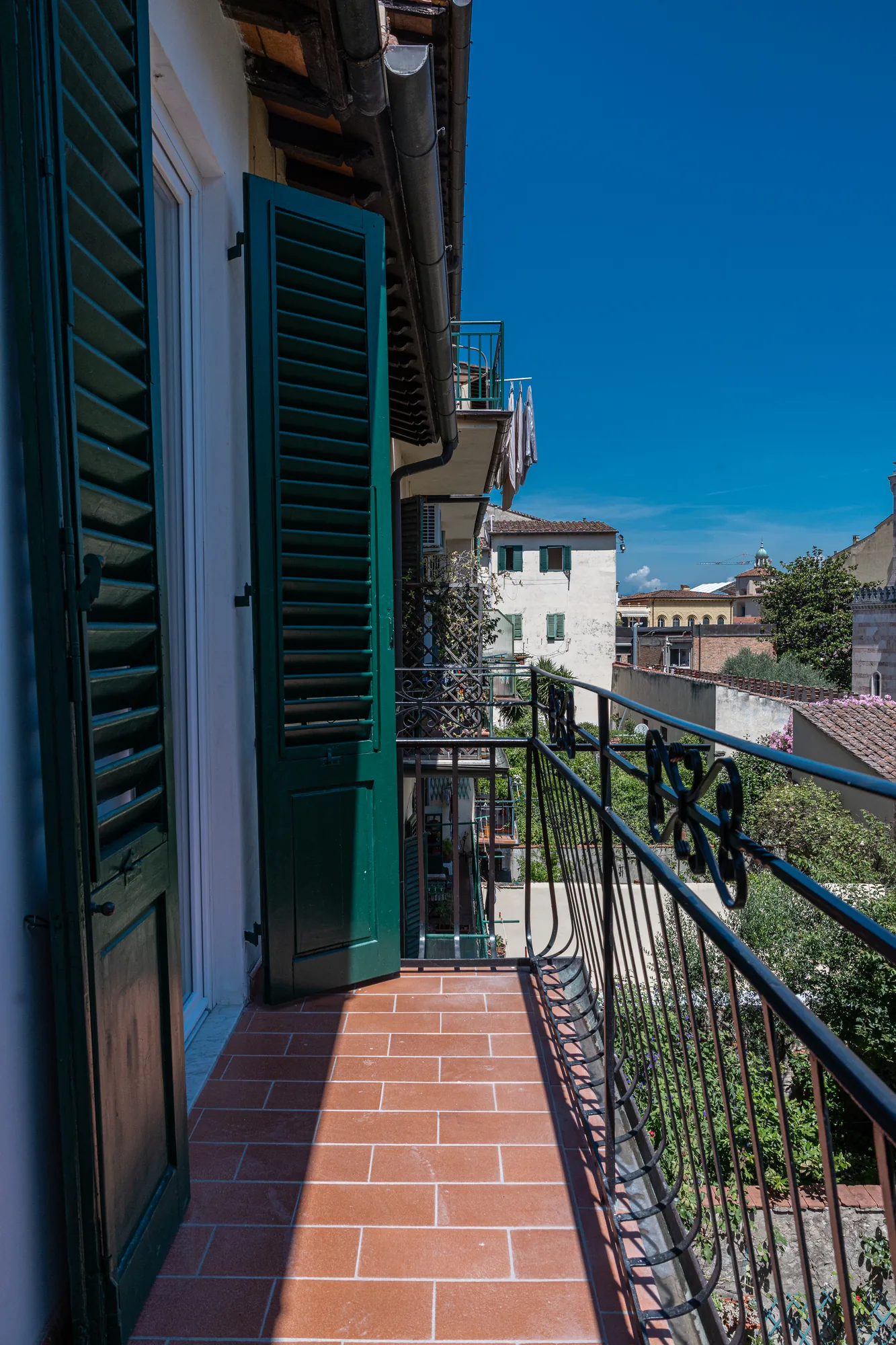 Balcony with iron railing and green shutters, overlooking the city rooftops under a clear sky