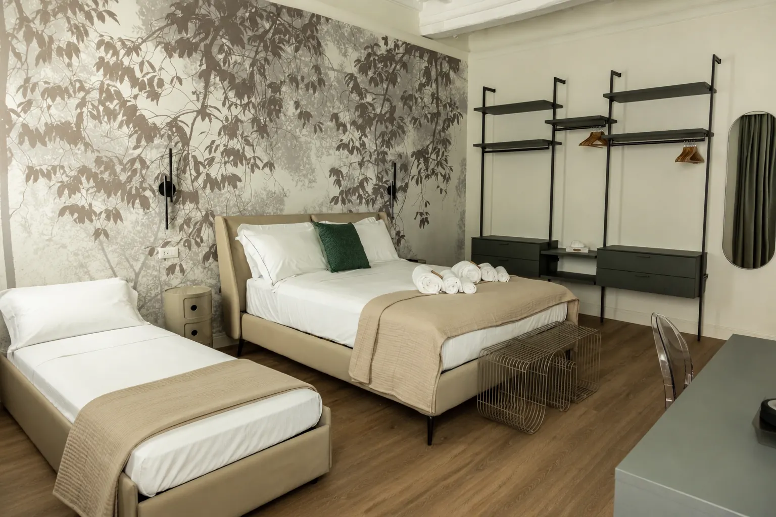 Camera da letto moderna con due letti, parete decorata con foglie, armadio e scrivania. Asciugamani arrotolati sul letto