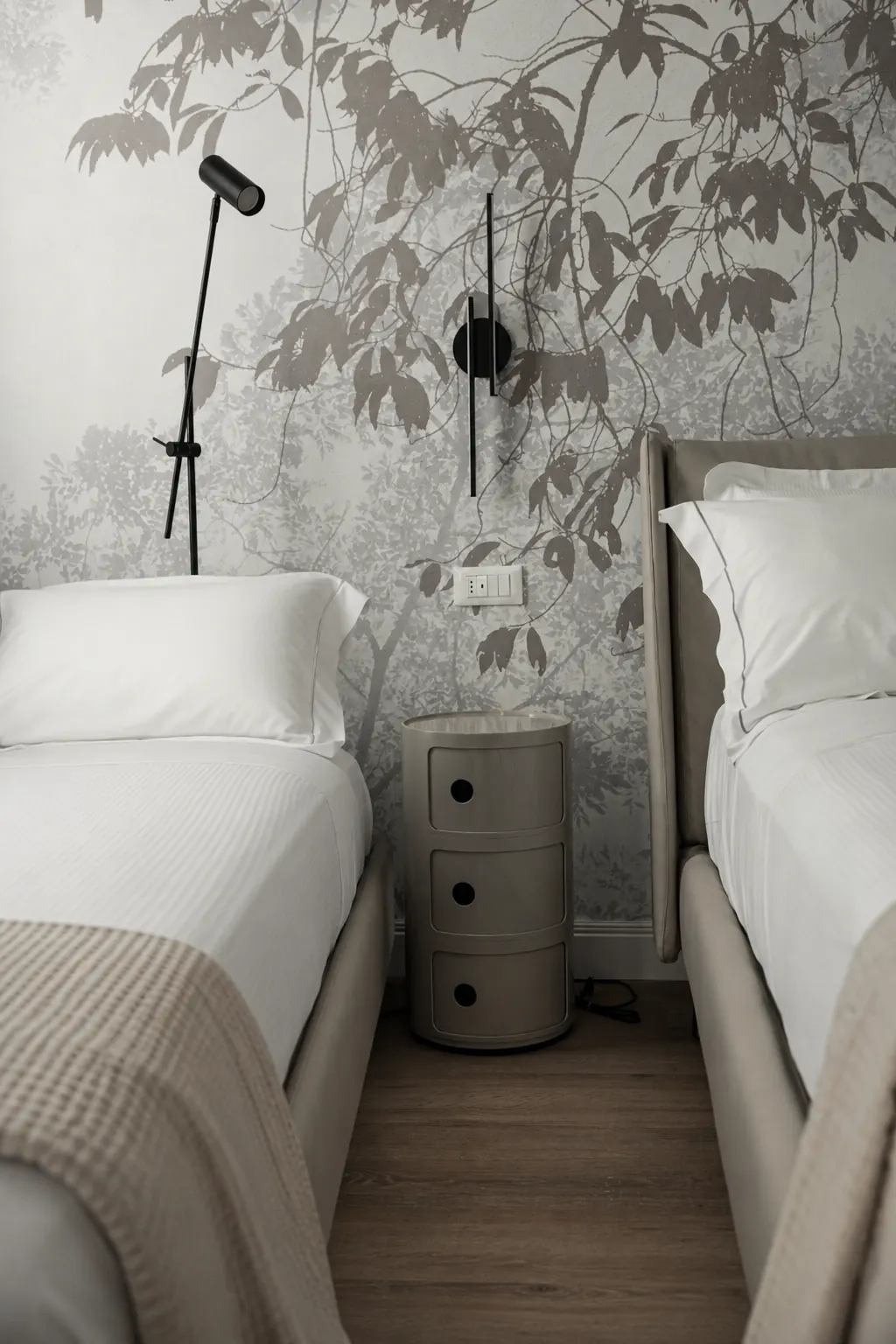 Due letti con testiere beige, un comodino cilindrico accanto e una parete decorata con foglie grigie