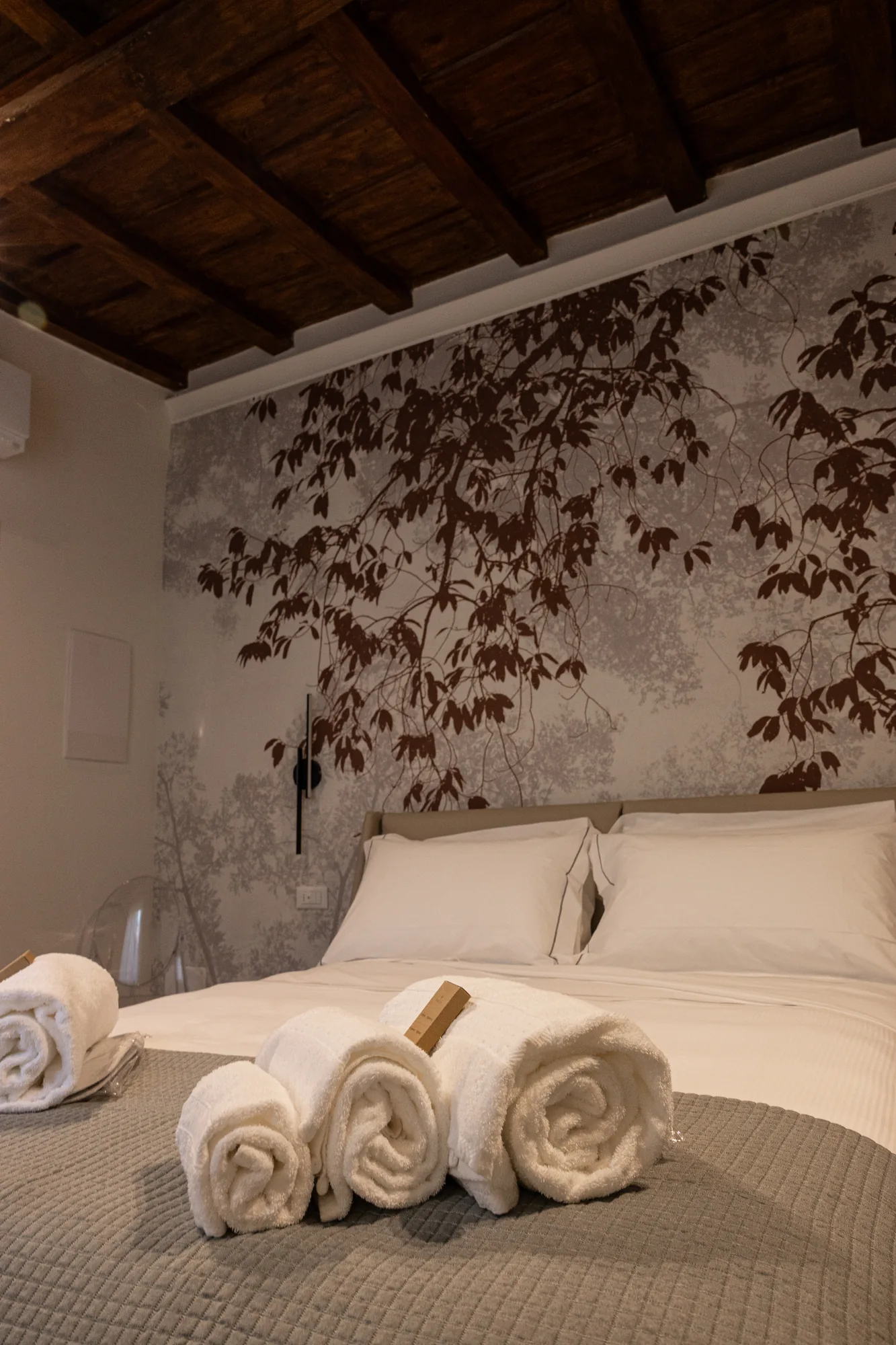 Bedroom with made bed, rolled towels, and wall decorated with a branch and leaf pattern