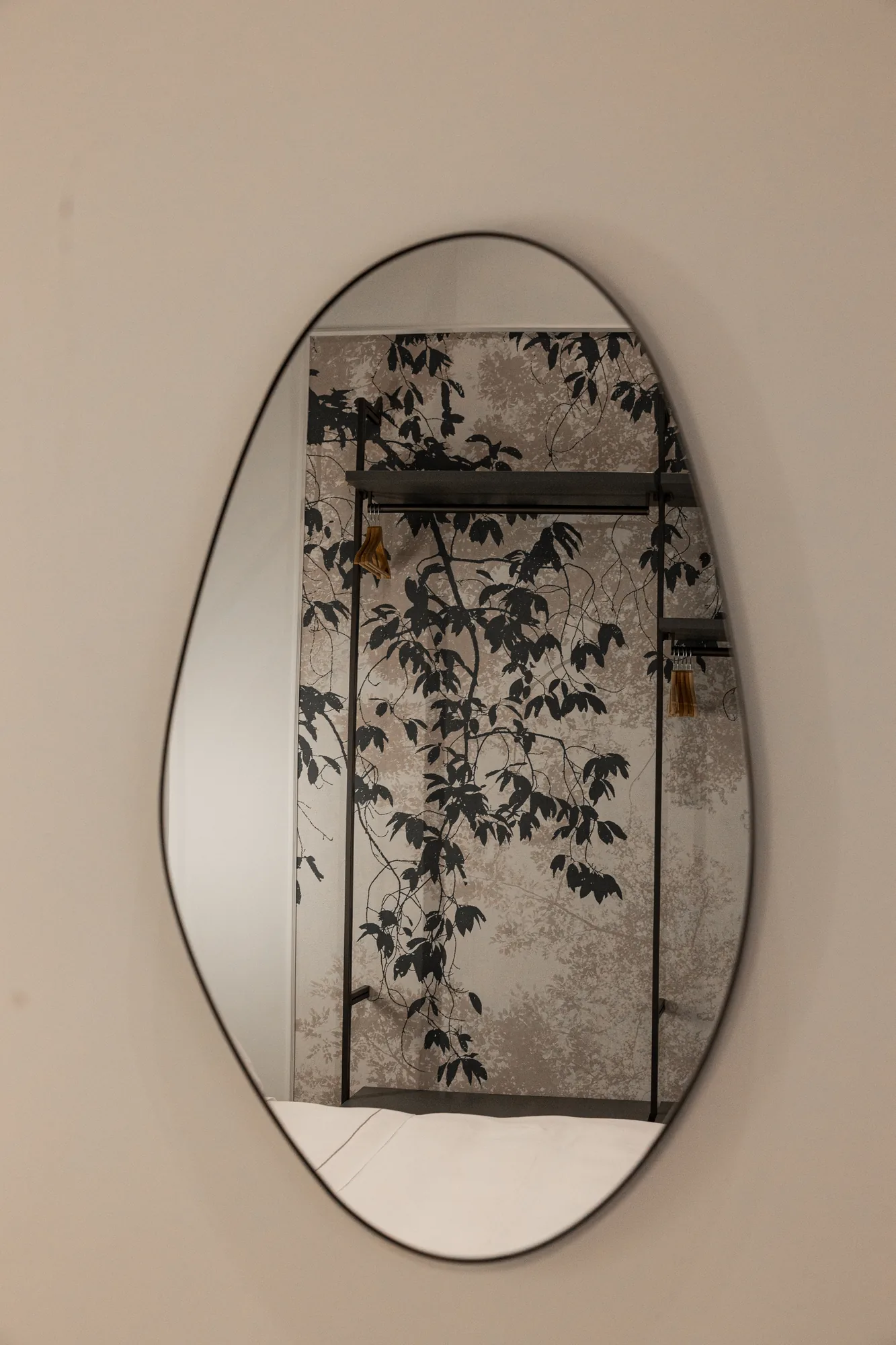 Oval mirror reflecting a wall with leaf-patterned wallpaper and a clothes rack