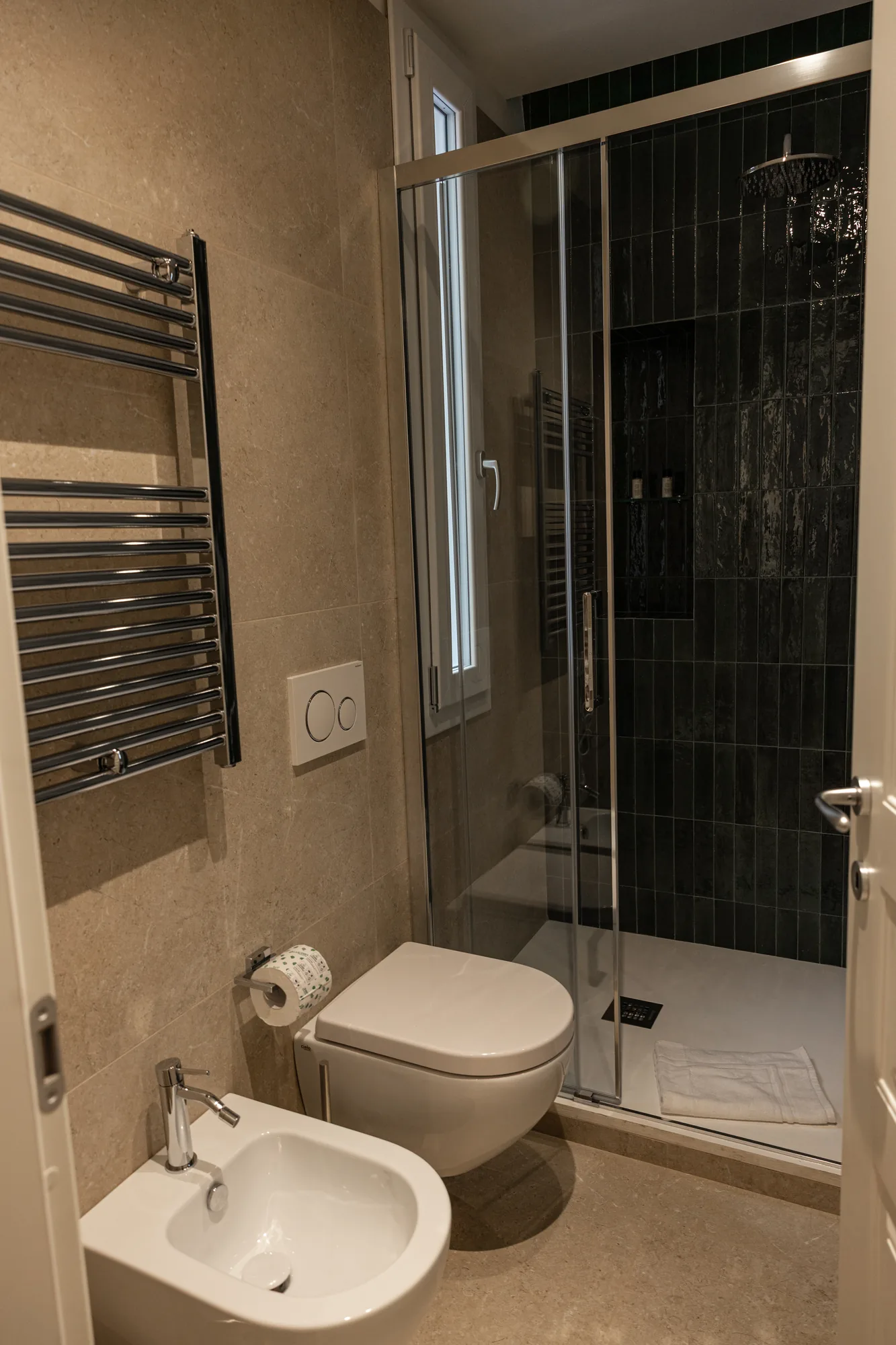 Modern bathroom with glass shower, toilet, and bidet, covered in beige stone and black tiles