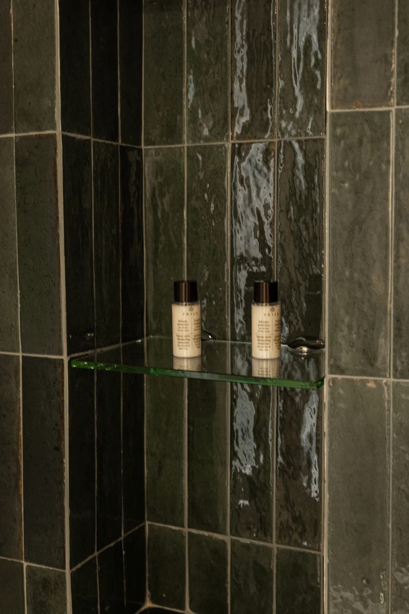 Two bottles of shower product on a glass shelf in a dark tiled niche